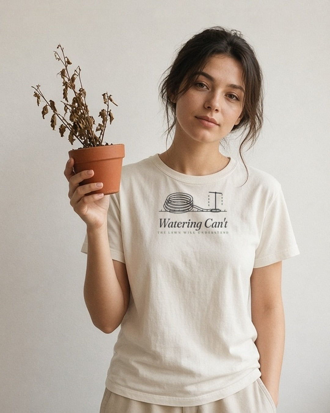 The Watering Can't Tee in Ivory, young woman against a plain wall holding up a pot of dried-up plants, other hand in her pocket