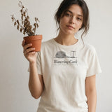 The Watering Can't Tee in Ivory, young woman against a plain wall holding up a pot of dried-up plants, other hand in her pocket