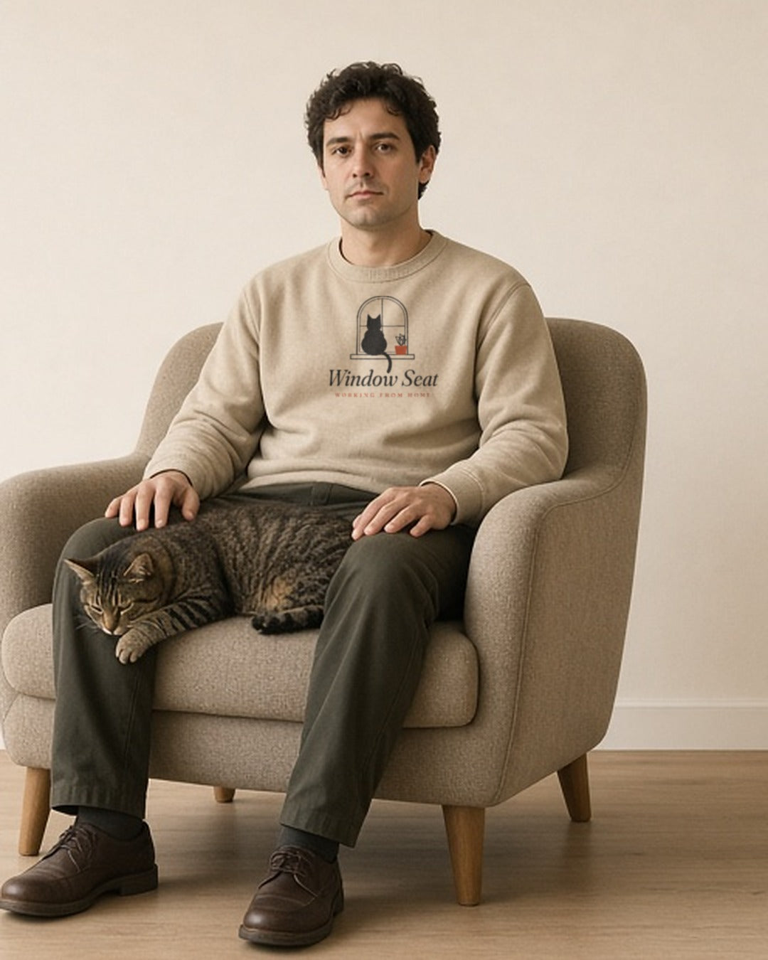The Window Seat Crewneck in Sand, man in the Sand crew in a cream armchair, a tabby cat asleep on his lap, pale wall