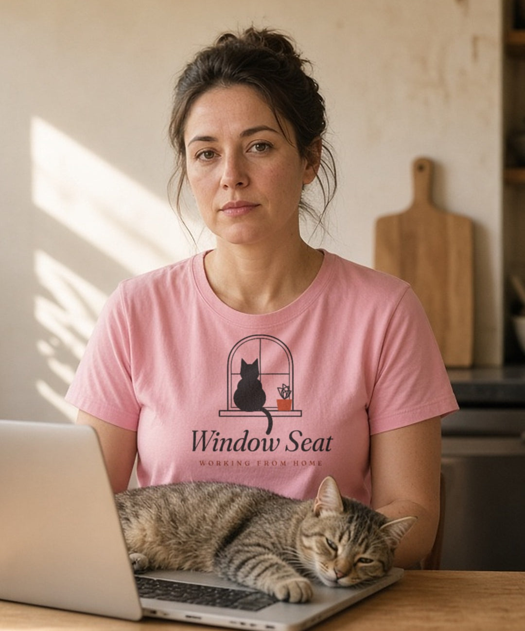 The Window Seat Tee in Blossom, a woman (30s) in the Blossom tee at a kitchen table with a laptop, while the real worker, a cat, occupies the sunny windowsill and the keyboard