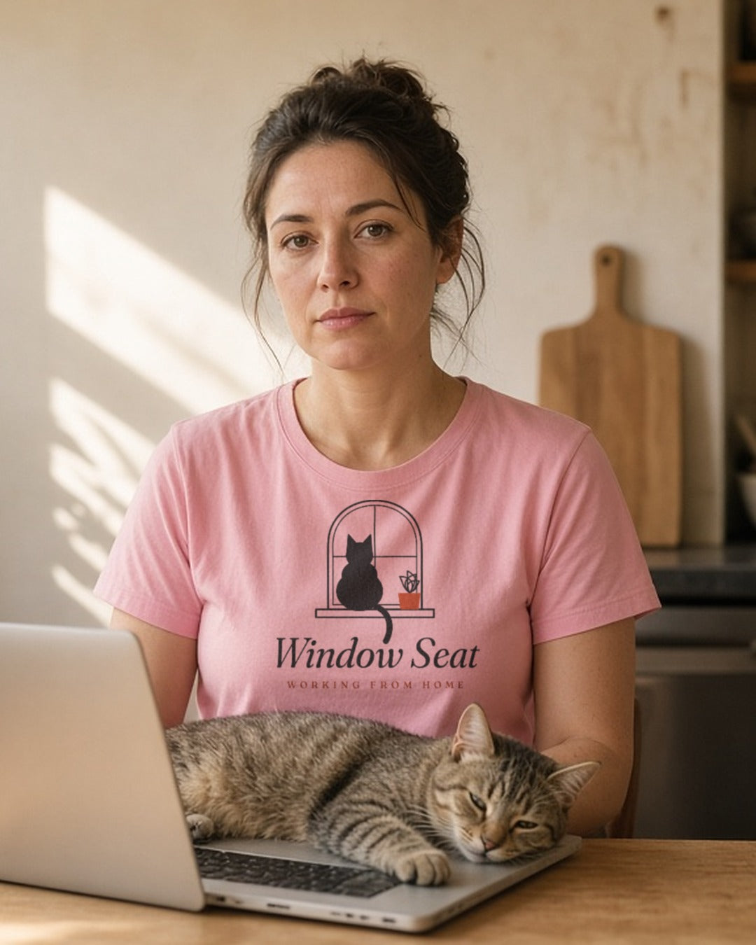 The Window Seat Tee in Blossom, a woman (30s) in the Blossom tee at a kitchen table with a laptop, while the real worker, a cat, occupies the sunny windowsill and the keyboard