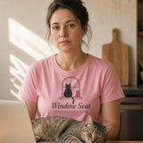 The Window Seat Tee in Blossom, a woman (30s) in the Blossom tee at a kitchen table with a laptop, while the real worker, a cat, occupies the sunny windowsill and the keyboard