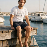 The Yacht Club Tee in White, a man (30s) in the White tee sits on a wooden dock with his feet over the water, holding a tiny toy sailboat on a string like it is his fleet